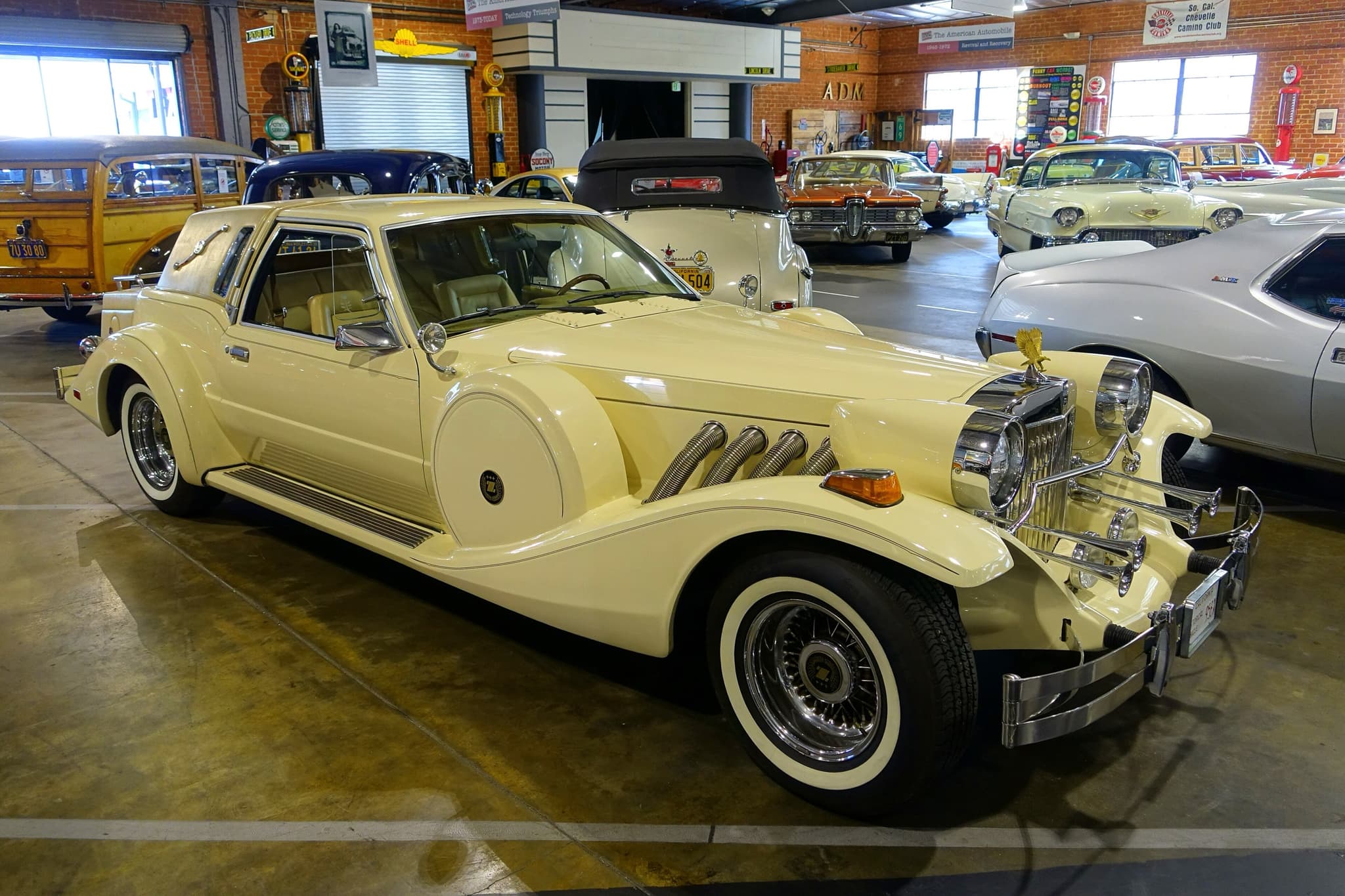 A pale yellow Golden Spirit coupé with a chrome grille, gold eagle, two large headlamps, fog lamps, trumpet horns and flared wings over white-wall wire wheels, in a museum hall among other cars.